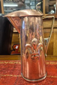 a copper jug with lid and embellished fleur de lys design.