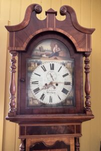A period long case clock often referred to as a Grandfather clock with a painted dial with folk scenes and a Wrexham makers mark